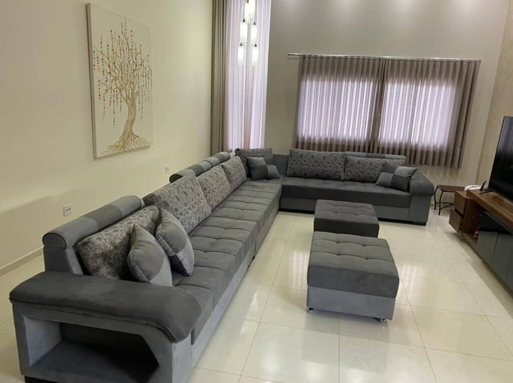 Reforma de sofá gigante em formato de L com pufes complementares em tecido cinza. Serviço de estofaria de alto padrão e renovação de móveis grandes, realizado em uma ampla sala de estar moderna na Barra da Tijuca, Rio de Janeiro.